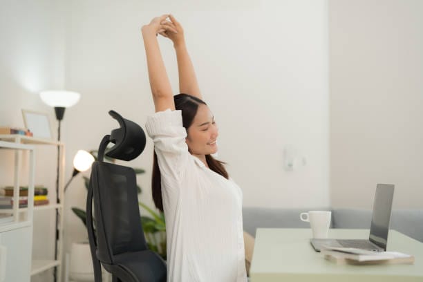 Asian woman in a white blouse takes a refreshing stretch break in her minimalist home office setup, with a laptop, coffee cup, and a bright, airy background enhancing her relaxed posture