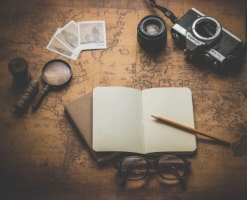 Vintage treasure map background with an open notebook, pencil, camera, magnifying glass, photographs, eyeglasses, and twine arranged on a desk, evoking exploration and adventure